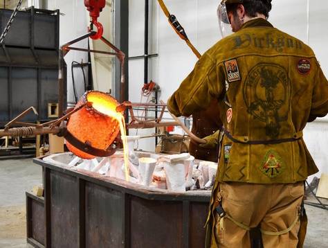 Man in working gear overlooking a smoldering bin of molten metal pouring into a mold.