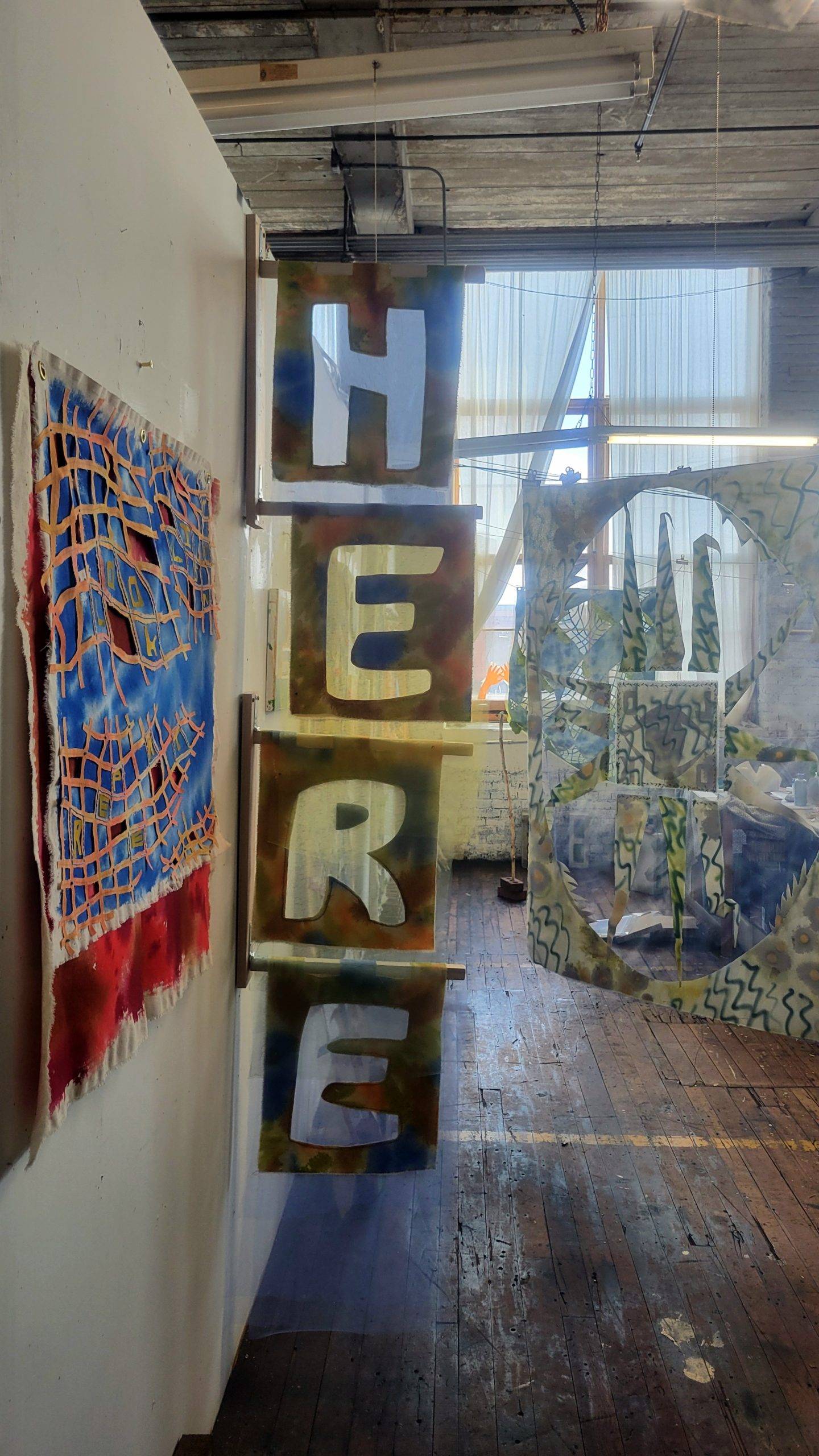 Photograph inside an artist's studio with a sculptural installation of floating letters reading "Here".