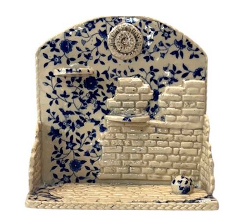A small ceramic work depicting a portion of a room with partial brick wall and floral wallpaper.