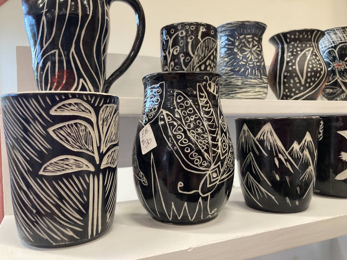 Photograph of two rows of ceramic vases and pitchers, black hued with illustrative white designs of natural objects like plants, butterflies, and mountains.