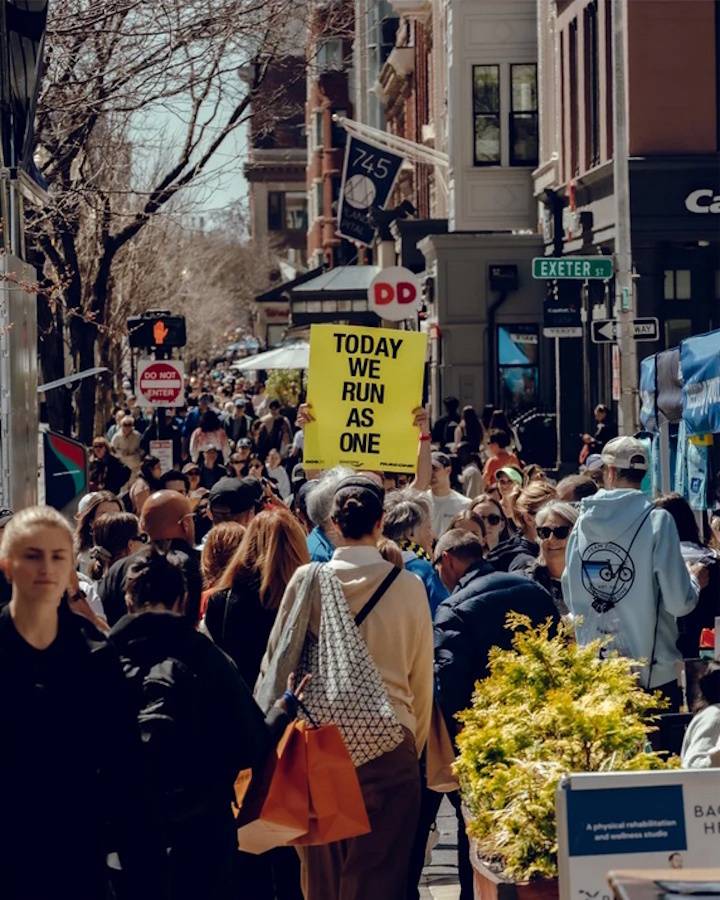 A photograph of a densely packed crowd of spectators at the Boston Marathon, with one onlooker holding a sign saying "Today we run as one."